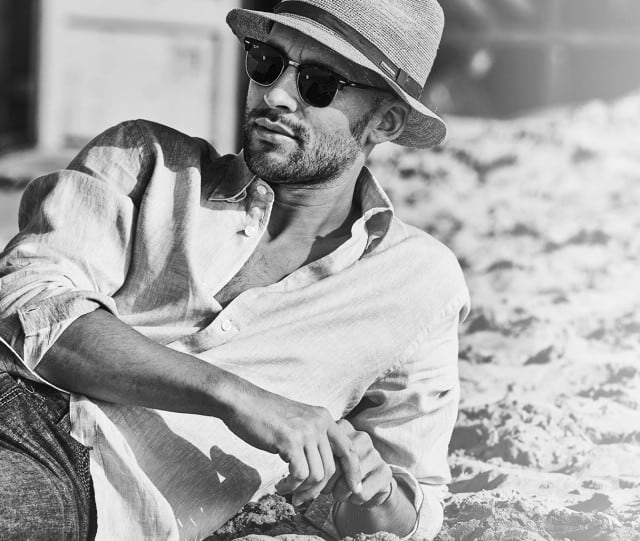 Eine Person mit Sonnenbrille und Hut liegt lässig im Sand und trägt ein helles Leinenhemd sowie dunkle Jeans in einer kontrastreichen Schwarz-Weiß-Aufnahme.