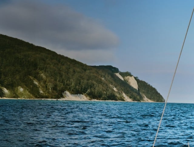 Auf dem Bild ist ein Mann auf einem Segelboot zu sehen, der ein rosa Poloshirt, eine grüne Hose und eine Armbanduhr trägt.