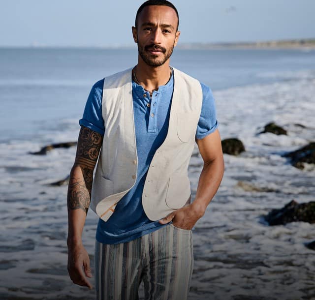 Eine Person in einem blauen T-Shirt, einer hellen Weste und gestreiften Hosen steht an einer felsigen Küste direkt am Wasser.