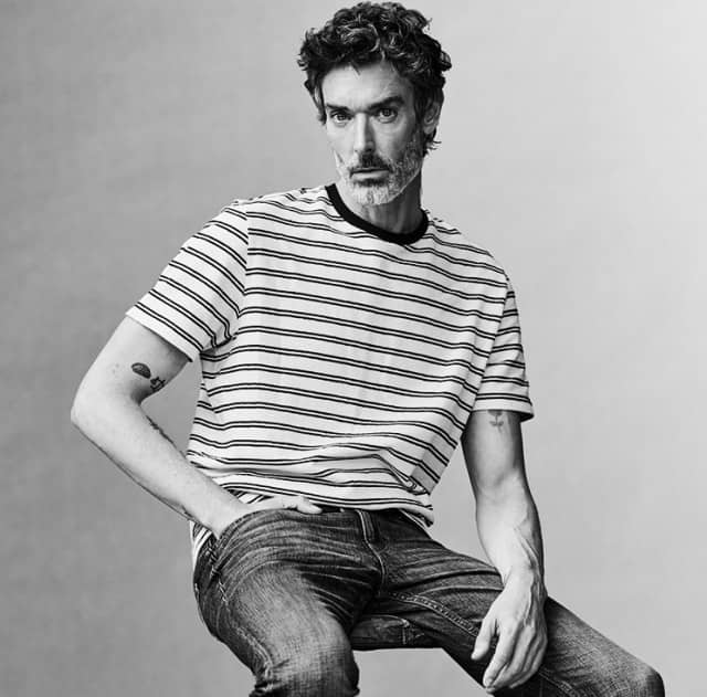 Eine Person trägt ein gestreiftes T‑Shirt mit dunklem Rundhalsausschnitt und eine dunkle Jeans und sitzt vor einem schlichten Studiohintergrund.