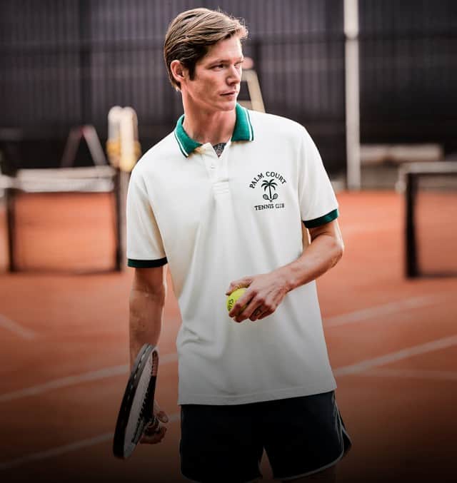Eine Person in einem weißen Poloshirt und dunklen Shorts steht mit Tennisschläger und Tennisball auf einem Tennisplatz.