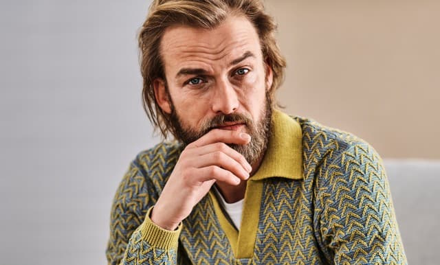 Mann im gemusterten blau-gelben Strickcardigan mit gelbem Kragen und weißem Shirt. Retro-inspirierter, auffälliger Stil.
