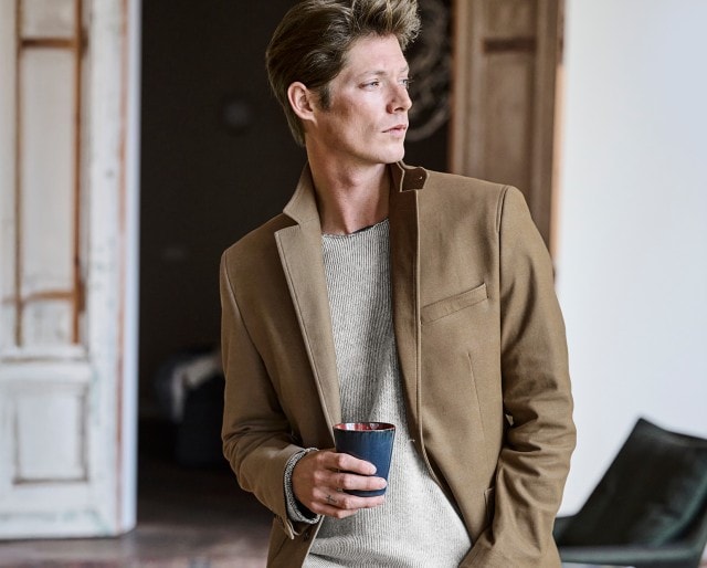 Auf dem Bild ist ein Mann zu sehen, der einen beigen Blazer über einem hellen Strickpullover trägt und in entspannter Haltung eine Tasse in der Hand hält.
