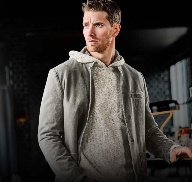 Eine Person trägt einen hellen Kapuzenpullover unter einer grauen Jacke und steht in einem modern gestalteten Innenraum.