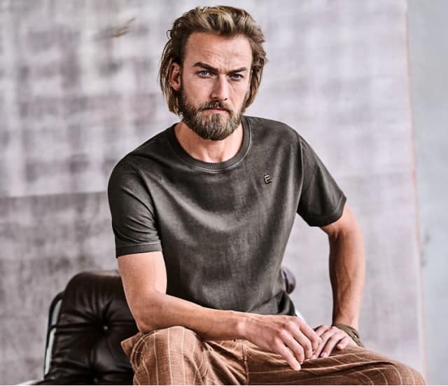 Eine Person trägt ein dunkles T‑Shirt und eine karierte Hose und sitzt auf einem Ledersessel vor einer schlichten Wand.