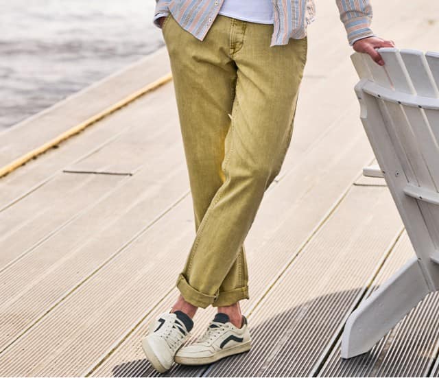 Eine Person trägt eine olivgrüne Hose, weiße Sneaker und ein gestreiftes Oberteil und steht auf einem Holzsteg am Wasser.