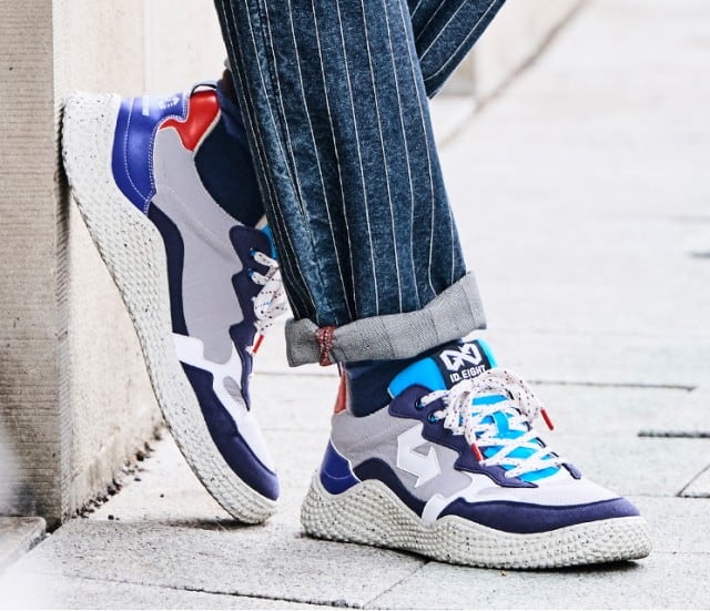 Ein Paar mehrfarbige Sneaker in Weiß, Blau und Rot wird zu einer dunkel gestreiften Hose getragen und vor einer hellen Steinwand präsentiert.