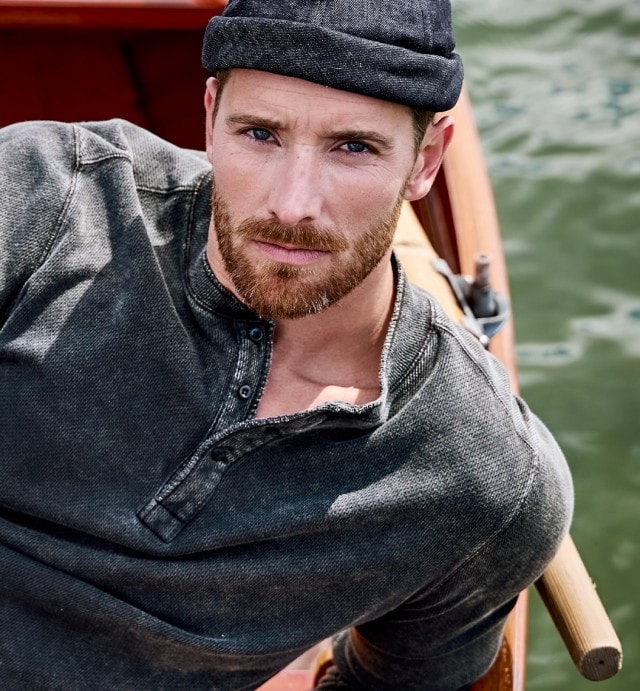 Auf dem Bild ist ein Mann zu sehen, der ein dunkelgraues Henley-Longsleeve, eine graue Hose und eine dunkle Mütze trägt, während er in einem Boot am Wasser sitzt.
