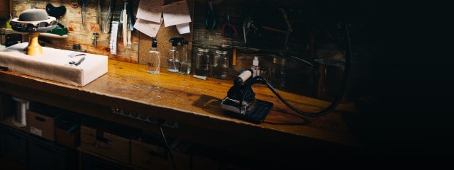 Auf dem Bild ist eine Werkbank zu sehen, auf der ein halbfertiger Filzhut auf einem Holzblock, eine Schere sowie ein Bügeleisen liegen, während an der Wand verschiedene Werkzeuge hängen.