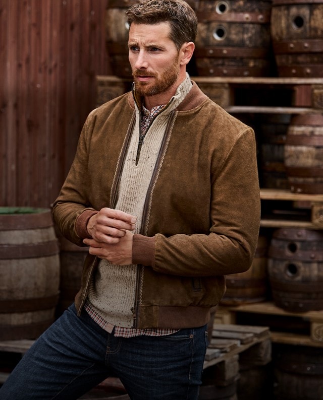Ein Mann trägt eine braune Wildlederjacke über einem beigen Strickpullover mit Reißverschluss, darunter ein kariertes Hemd und kombiniert das Outfit mit einer dunklen Jeans.