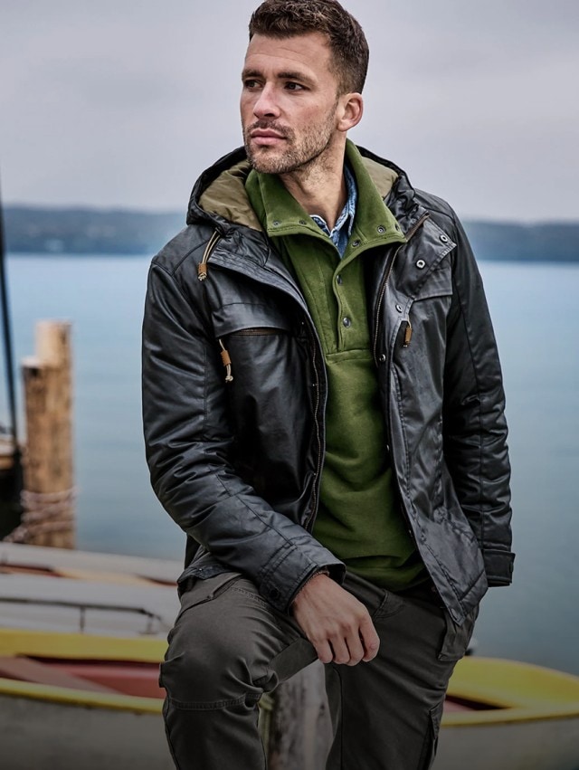 Ein Mann trägt eine schwarze Lederjacke über einem grünen Pullover mit Knopfleiste und einem blauen Hemd, kombiniert mit einer grauen Hose.