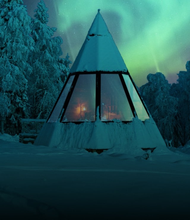 Ein schneebedecktes Zelt mit warmem Licht im Inneren steht inmitten eines winterlichen Waldes, während darüber eindrucksvoll die Nordlichter am Himmel leuchten.