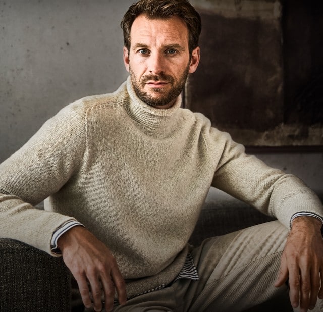 Ein Mann trägt einen beigefarbenen Strickpullover mit hohem Kragen über einem gestreiften Hemd und einer hellen Hose.