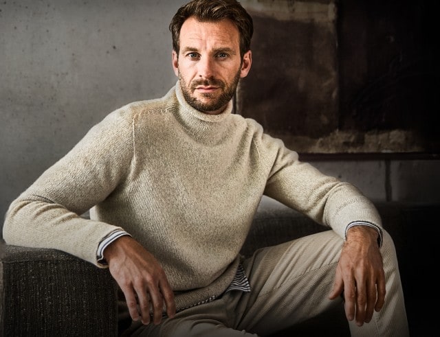 Ein Mann trägt einen weißen Strickpullover mit hohem Kragen und lehnt entspannt an einer grauen Wand.