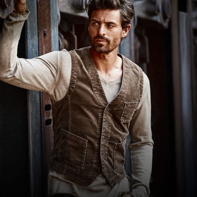 Auf dem Bild ist ein Mann zu sehen, der lässig an einem alten Zug oder Güterwaggon lehnt und dabei eine braune Weste über einem beigen Langarmshirt trägt.