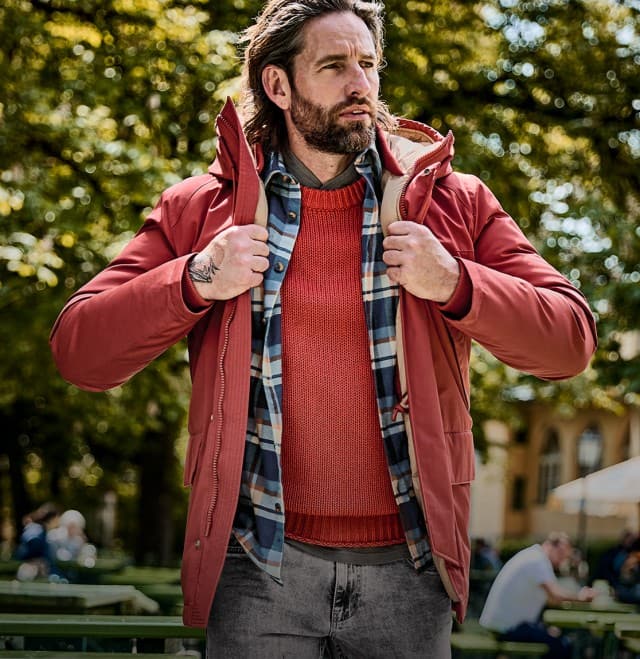 Auf dem Bild ist ein Mann zu sehen, der im Freien steht und eine rote Jacke über einem roten Pullover und einem karierten Hemd trägt