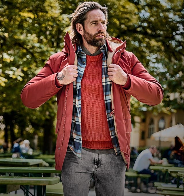 Ein Mann steht im Freien und trägt eine rote Winterjacke über einem rot-orangen Pullover und einem blau-weiß karierten Hemd, kombiniert mit grauen Jeans.