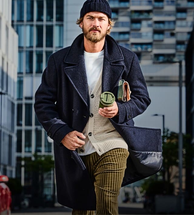 Der Mann trägt einen dunkelblauen Mantel über einem hellen Strickpullover mit weißem Shirt, kombiniert mit braunen Cordhosen und einer dunkelblauen Mütze für einen lässig-eleganten Herbstlook.