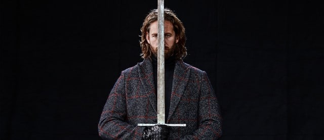 Auf dem Bild ist ein Mann mit langem Haar und Bart zu sehen, der in dunkler Kleidung und Handschuhen eine große zweischneidige Schwertklinge senkrecht vor seinem Gesicht hält. Auf dem Bild ist ein Mann mit langem Haar und Bart zu sehen, der in dunkler Kleidung und Handschuhen eine große zweischneidige Schwertklinge senkrecht vor seinem Gesicht hält.