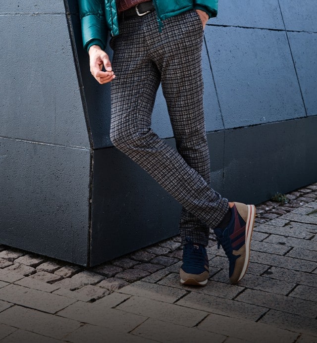 Auf dem Bild lehnt eine Person lässig an einer grauen Wand, trägt karierte Hosen, blaue Sneaker und eine grüne Jacke.