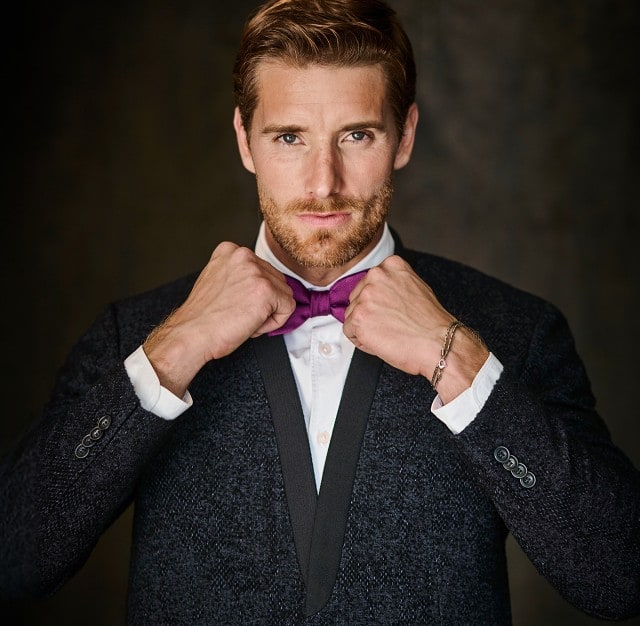 Auf dem Bild ist ein elegant gekleideter Mann zu sehen, der eine lila Fliege richtet und direkt in die Kamera blickt, vor einem dunklen, neutralen Hintergrund.