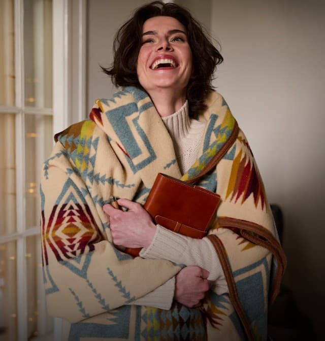 Auf dem Bild ist eine lachende Frau zu sehen, die sich in eine bunte, gemusterte Decke gehüllt hat und ein braunes Buch oder Notizbuch im Arm hält, während sie in einem gemütlich wirkenden Innenraum steht.