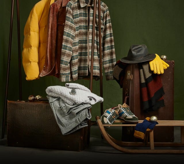 Auf dem Bild sind verschiedene Kleidungsstücke und Accessoires, darunter Jacken, Pullover, Schuhe, ein Hut, Handschuhe und ein Schal, dekorativ auf Koffern und einem Holzschlitten arrangiert, vor einem dunkelgrünen Hintergrund zu sehen.