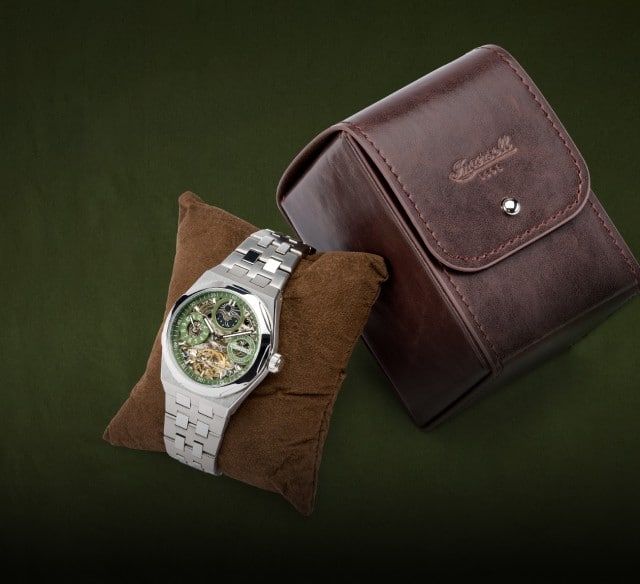 Auf dem Bild ist eine silberne Armbanduhr mit grünem Zifferblatt zu sehen, die auf einem braunen Kissen präsentiert wird, daneben liegt ein braunes Lederetui vor einem dunkelgrünen Hintergrund.