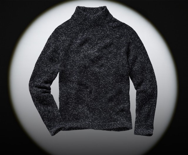 Auf dem Bild ist ein dunkelgrauer, grob gestrickter Pullover mit hohem Kragen zu sehen, der vor einem schwarz-weißen Hintergrund präsentiert wird.