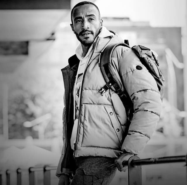 Ein Mann steht draußen in einer urbanen Umgebung an einem Geländer, trägt eine dicke Winterjacke und einen Rucksack und blickt nach vorne.