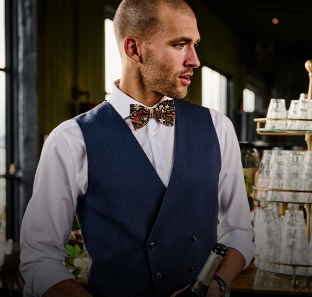 Ein elegant gekleideter Barkeeper schenkt in einer stilvollen Bar ein Getränk ein und blickt dabei zur Seite.