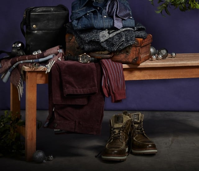 Auf dem Bild steht eine Holzbank, auf der ordentlich gefaltete Kleidung, Accessoires und ein paar dekorative Kugeln liegen, während darunter ein Paar braune Stiefel platziert ist.