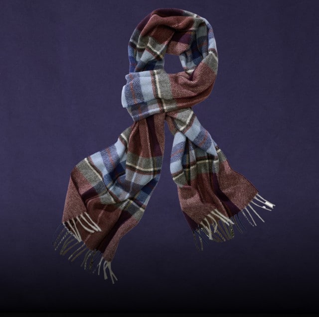 Auf dem Bild ist ein gemusterter Schal in Blau-, Grau- und Brauntönen zu sehen, der vor dunkelviolettem Hintergrund hängt.