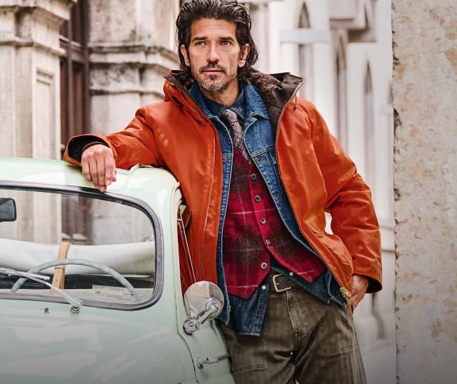 Ein Mann lehnt lässig an einem hellgrünen Oldtimer und trägt mehrere Schichten casualer Kleidung vor einer städtischen Steinfassade.