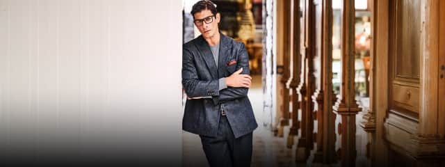 Ein Mann in einem dunkelgrauen Anzug steht mit verschränkten Armen lässig in einem hellen Gang mit holzverkleideten Säulen.