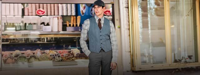 Ein Mann steht vor einer Eistheke und trägt eine Schirmmütze, ein kariertes Hemd, eine blaue Weste und eine graue Hose.