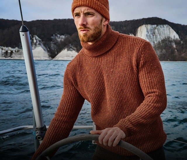 Ein Mann in einem rostroten Strickpullover und passender Mütze steuert auf einem Segelboot über das Meer, während im Hintergrund hohe, helle Kreidefelsen zu sehen sind.