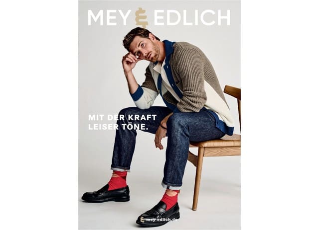 Auf dem Bild ist das Cover des Mey & Edlich Katalogs zu sehen aus dem ein Mann mit einer beigefarbenen Strickjacke in einer nachdenklichen Pose auf einem Stuhl sitzt.