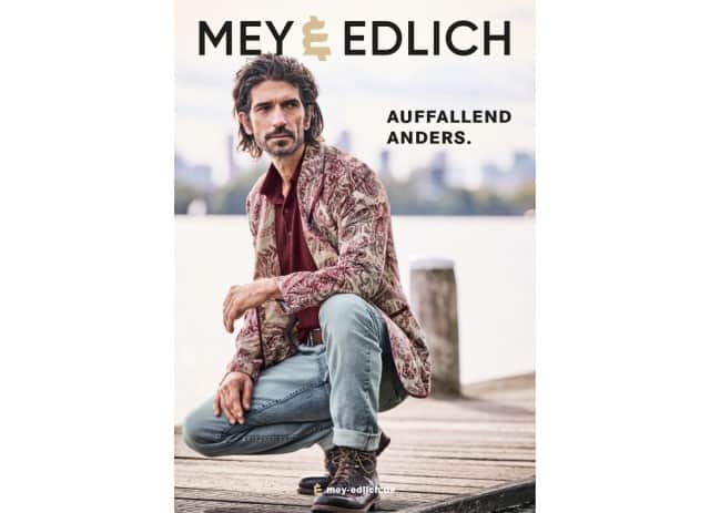 Auf dem Cover zu sehen ist ein Mann, der ein burgunderrotes Shirt unter einem beigefarbenen Mantel mit bordeauxrotem Paisley‑Muster sowie eine hellblaue Jeans und braune Boots trägt.