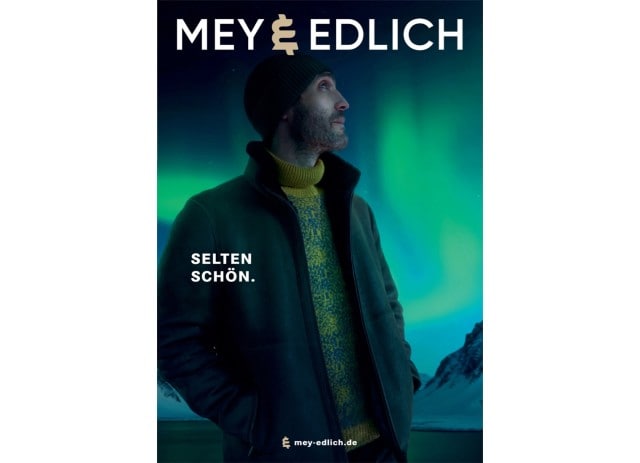 Ein Mann in winterlicher Kleidung steht vor einer Kulisse aus Polarlichtern und Bergen, während darüber der Markenname Mey & Edlich und der Slogan „Selten schön.“ prangen.