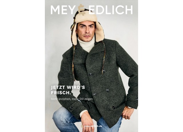 Das Cover zeigt einen Mann, der einen dunkelgrauen, doppelreihigen Wollmantel über einem weißen Rollkragenpullover trägt, kombiniert mit einer warm gefütterten Fellmütze und blauen Jeans.
