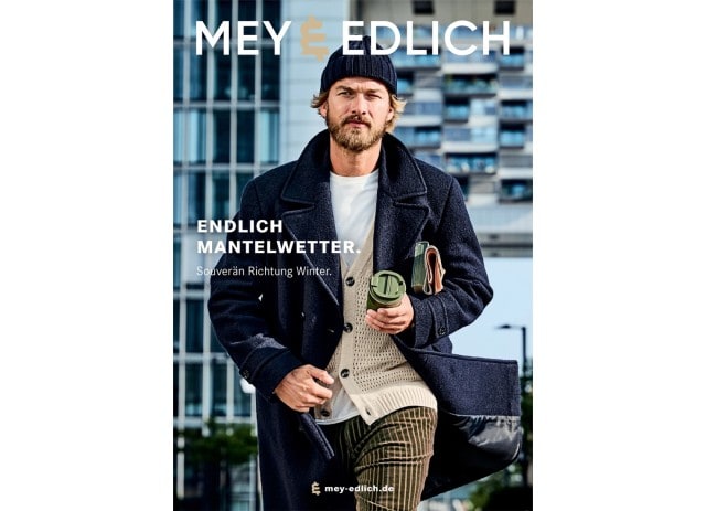 Ein Mann trägt einen dunkelblauen Wollmantel über einer grob gestrickten, cremefarbenen Strickjacke mit weißem T-Shirt, kombiniert mit einer dunkelblauen Strickmütze und braun gestreiften Cordhosen, während er eine Thermotasse und ein Notizbuch in der Hand hält.