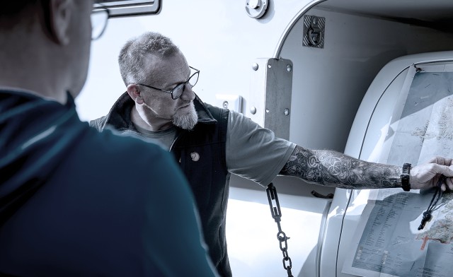Ein Mann mit Brille und tätowiertem Arm zeigt auf eine Landkarte, die an einem Fahrzeug befestigt ist, während eine weitere Person danebensteht und zuschaut.