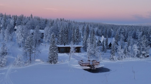 Eine verschneite Winterlandschaft mit schneebedeckten Bäumen, mehreren Hütten und einem Holzsteg, der zu einem Eisloch im zugefrorenen See führt, ist bei Abenddämmerung zu sehen.