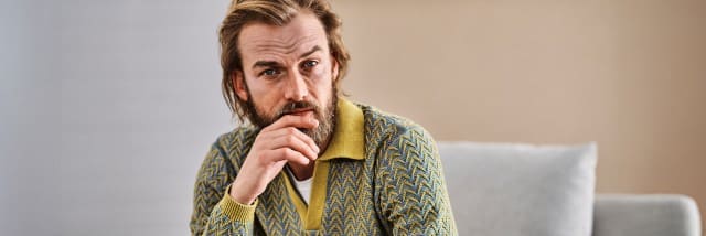 Mann im gemusterten blau-gelben Strickcardigan mit gelbem Kragen und weißem Shirt. Retro-inspirierter, auffälliger Stil.