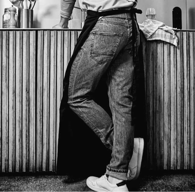 Lässige, graue Jeans mit aufgesetzten Taschen, kombiniert mit einem hellen Oberteil, dunkler Schürze und weißen Sneakern. Moderner Workwear-Stil.