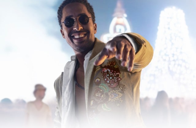 Ein Mann mit Sonnenbrille und bestickter Jacke steht im Vordergrund einer Menschenmenge und zeigt lächelnd in die Kamera, während im Hintergrund Lichter und eine große, helle Struktur sichtbar sind.