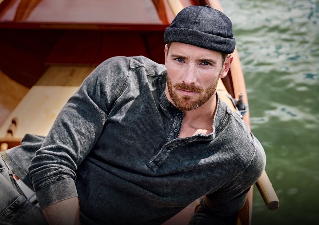 Ein Mann mit dunklem Henley-Shirt und Mütze liegt in einem Holzboot auf dem Wasser.