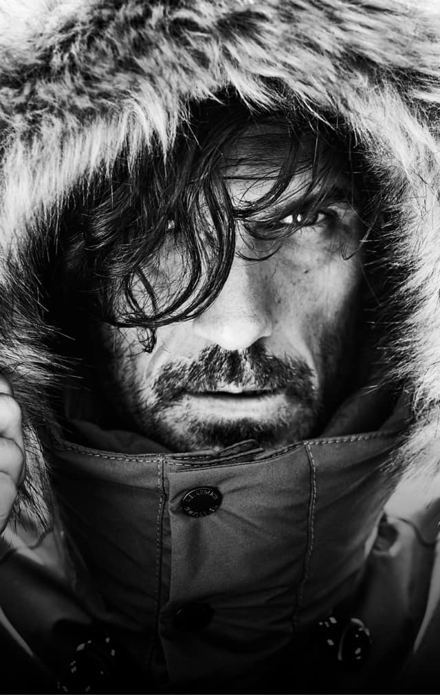 Eine Person trägt eine Winterjacke mit Pelzkapuze und hält diese mit beiden Händen fest um ihr Gesicht.