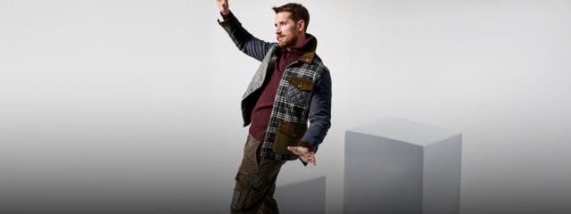 Eine Person trägt eine gemusterte Jacke mit verschiedenen Stoffeinsätzen, ein weinrotes Oberteil und braune Cargohosen und scheint im Gleichgewicht zu posieren.